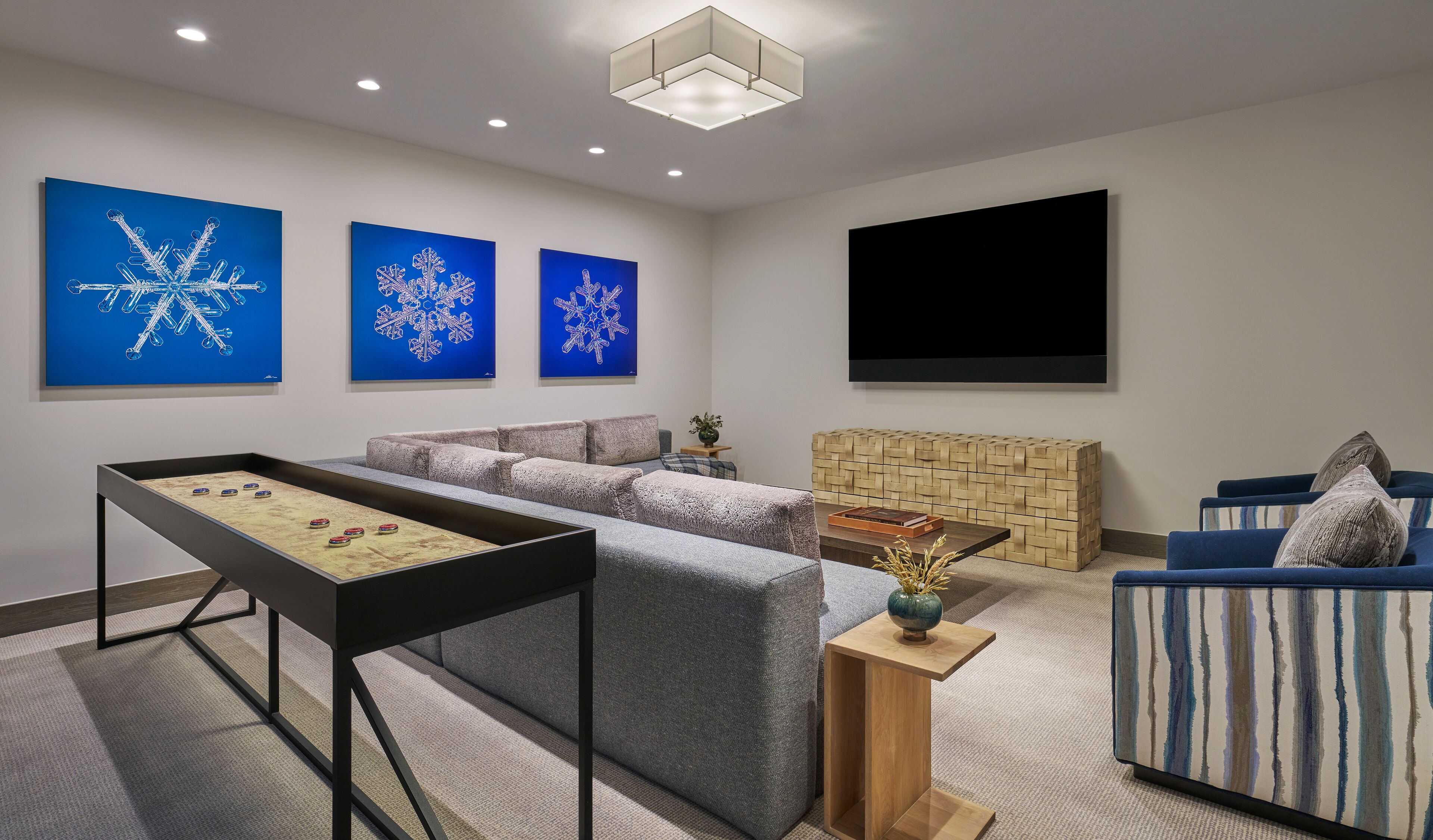 A modern living room with a foosball table, a large sofa, a television, and three blue snowflake paintings on the wall.