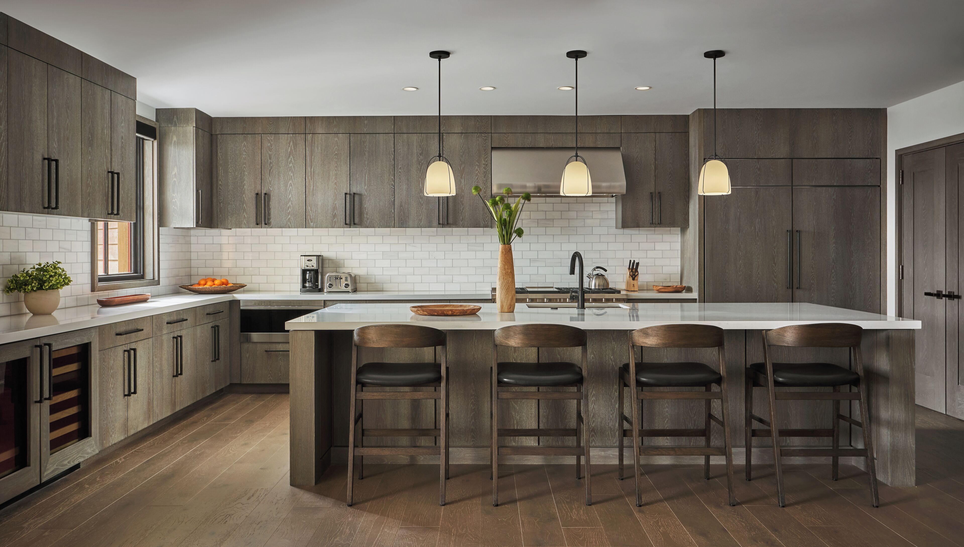 A modern kitchen with dark wood cabinets, a central island with bar stools, pendant lighting, and a white backsplash.