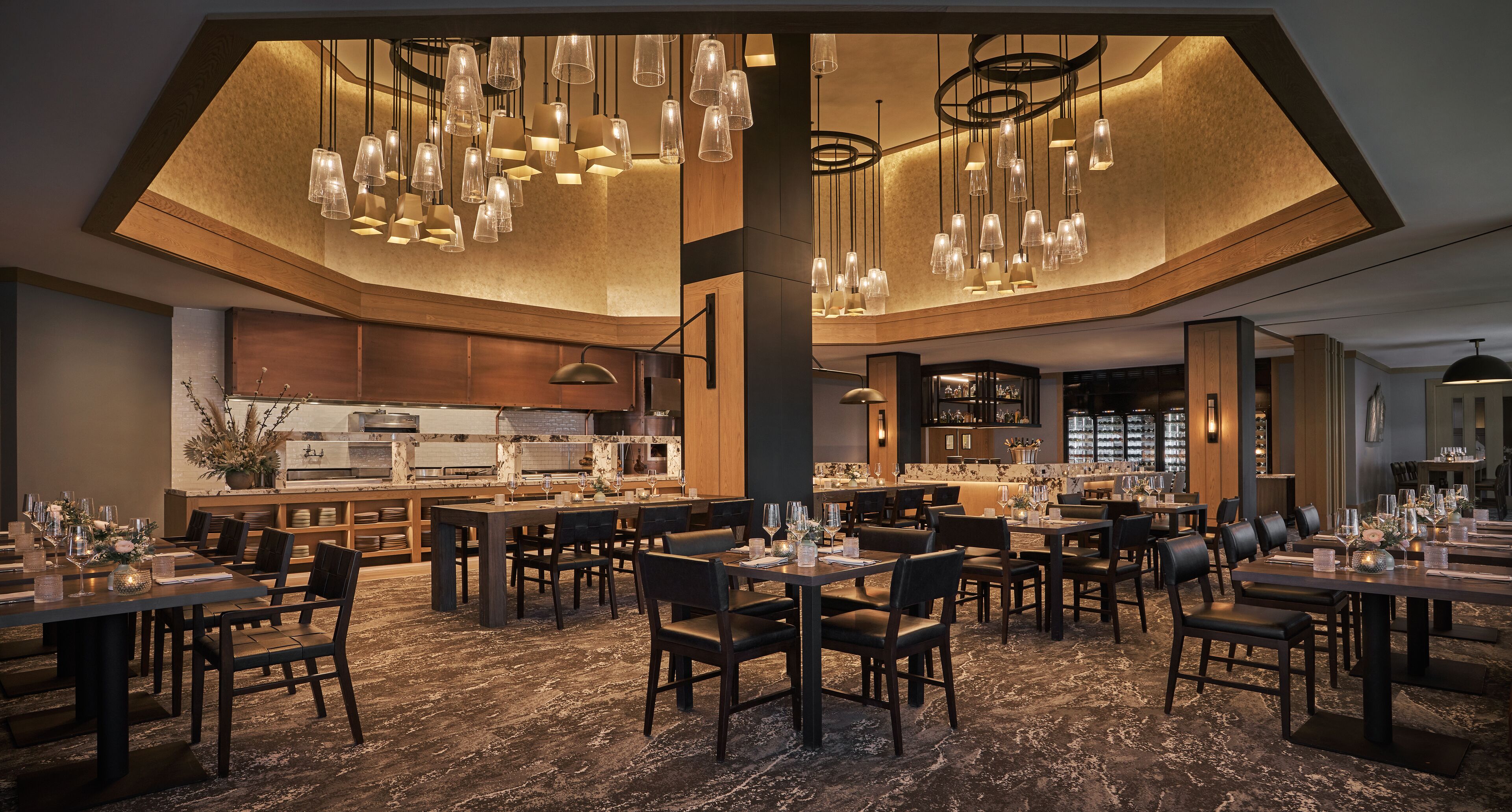A spacious restaurant with multiple dining tables and chairs, a central kitchen area, and modern pendant lighting.
