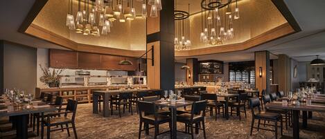 A spacious restaurant with multiple dining tables and chairs, a central kitchen area, and modern pendant lighting.