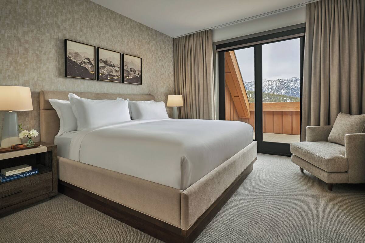 Ein Hotelzimmer mit einem großen Bett, einem Sessel, einem Nachttisch mit Lampe und Blick auf Berge durch das Fenster.