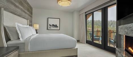 A modern hotel room with a large bed, a fireplace, a balcony with a view of the forest, and a framed artwork on the wall.