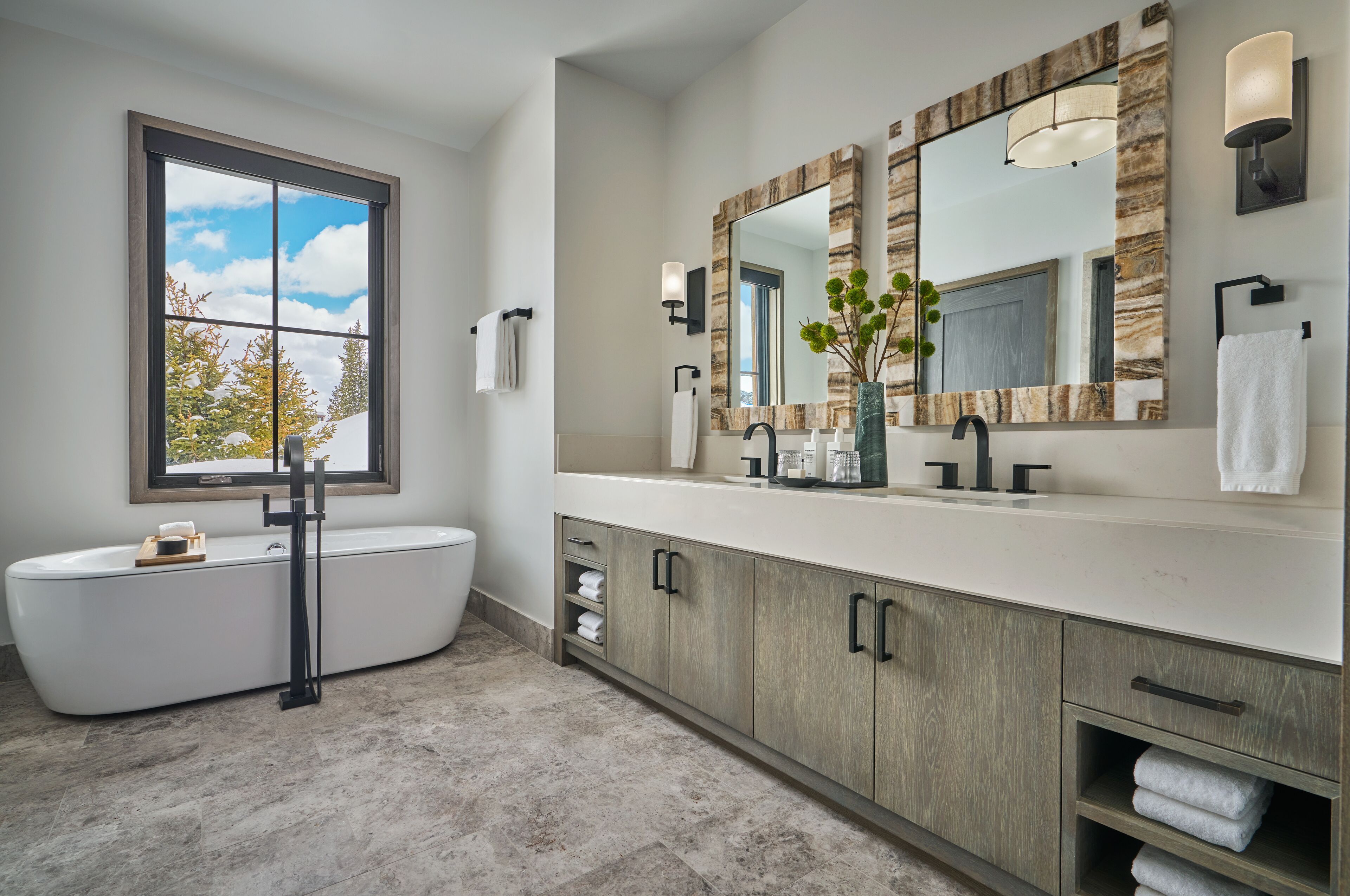 A modern bathroom with a freestanding bathtub, a large mirror with a wood frame, and a window with a view of trees.