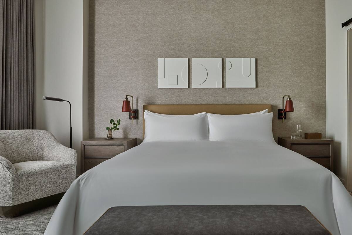 Une chambre d’hôtel moderne avec un grand lit, une chaise, des tables de chevet et des œuvres d’art accrochées au mur.