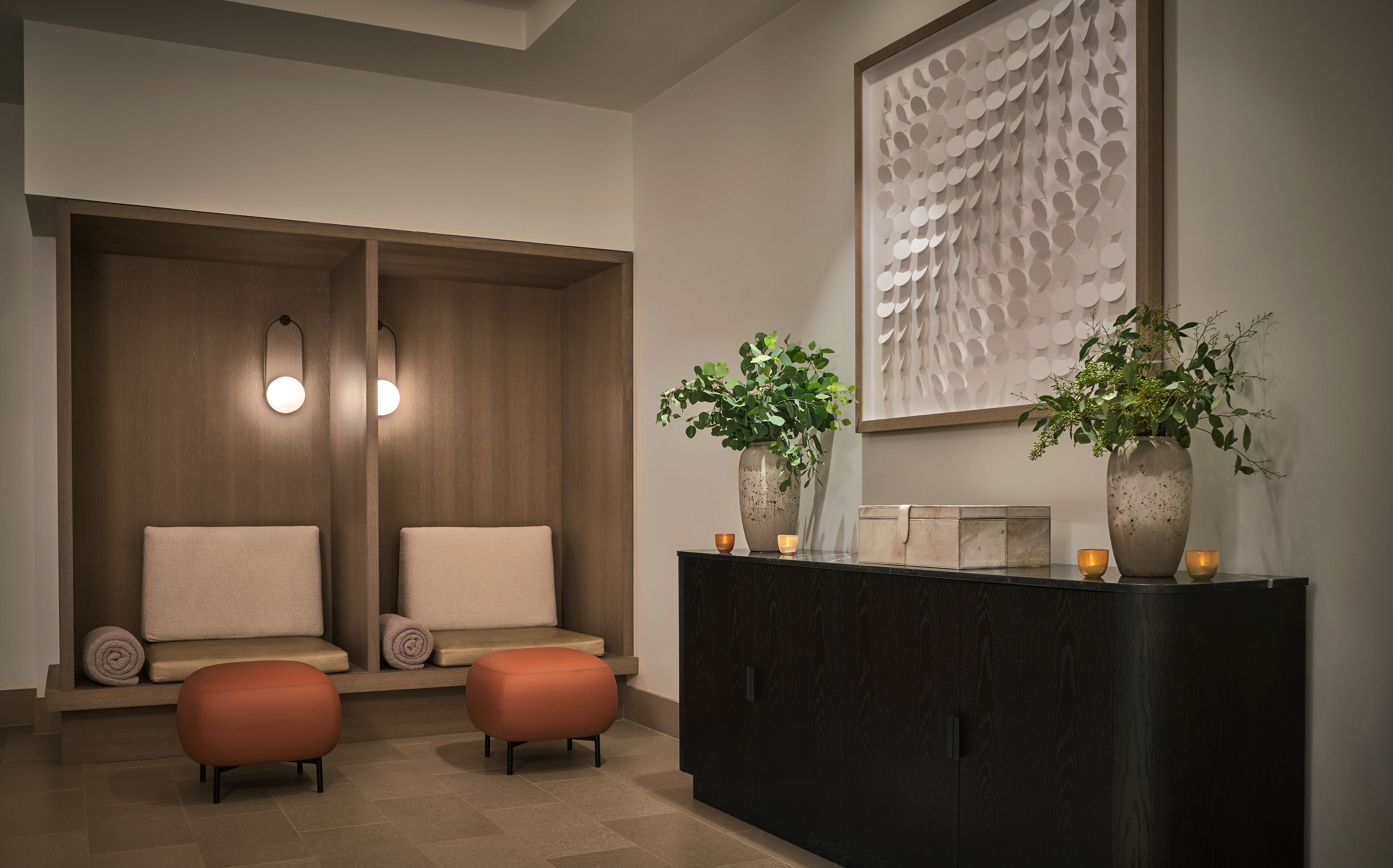 A modern spa room with a wooden bench, two ottomans, a dark wooden cabinet, and a wall-mounted artwork.
