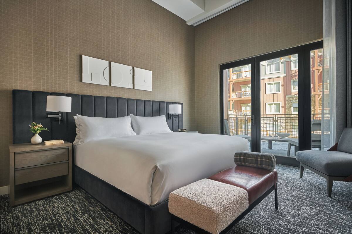 Une chambre d’hôtel avec un grand lit, deux tables de chevet avec des lampes, un banc et une vue sur la ville grâce à de grandes fenêtres.