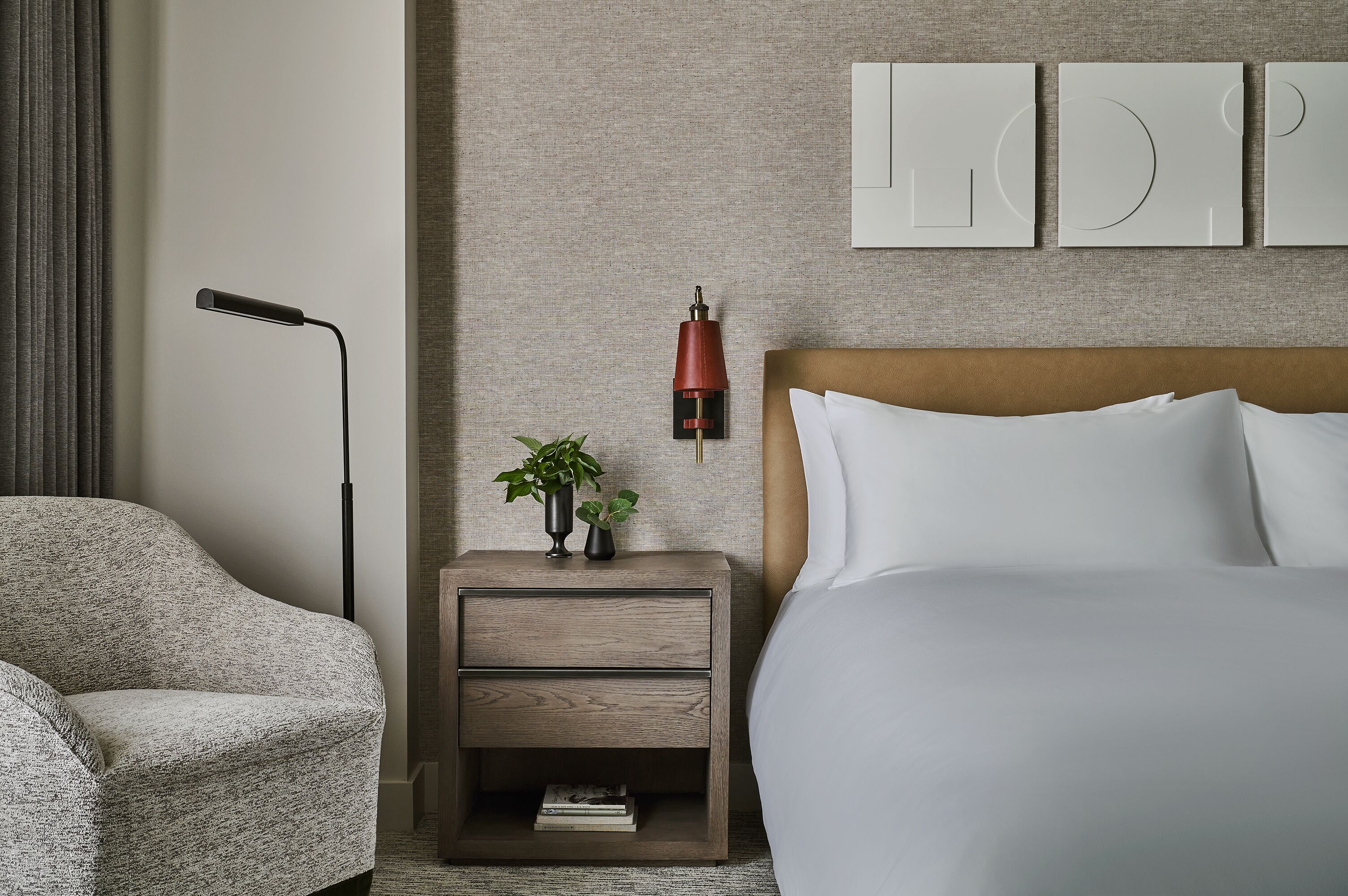 A modern hotel room with a bed, a nightstand with a lamp, a chair, and a wall with framed artwork.