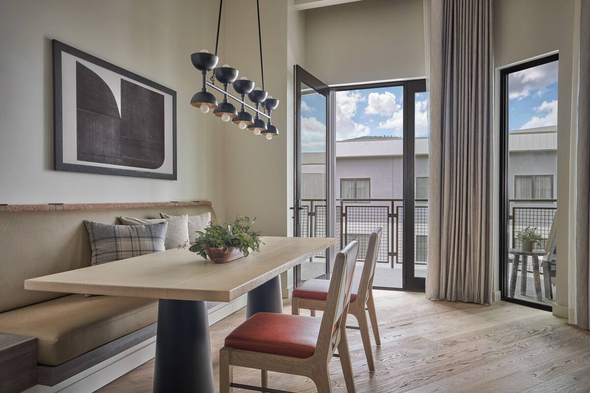 Un salon moderne comprenant un canapé, une table à manger, des chaises, un luminaire suspendu et une grande fenêtre donnant sur des immeubles.