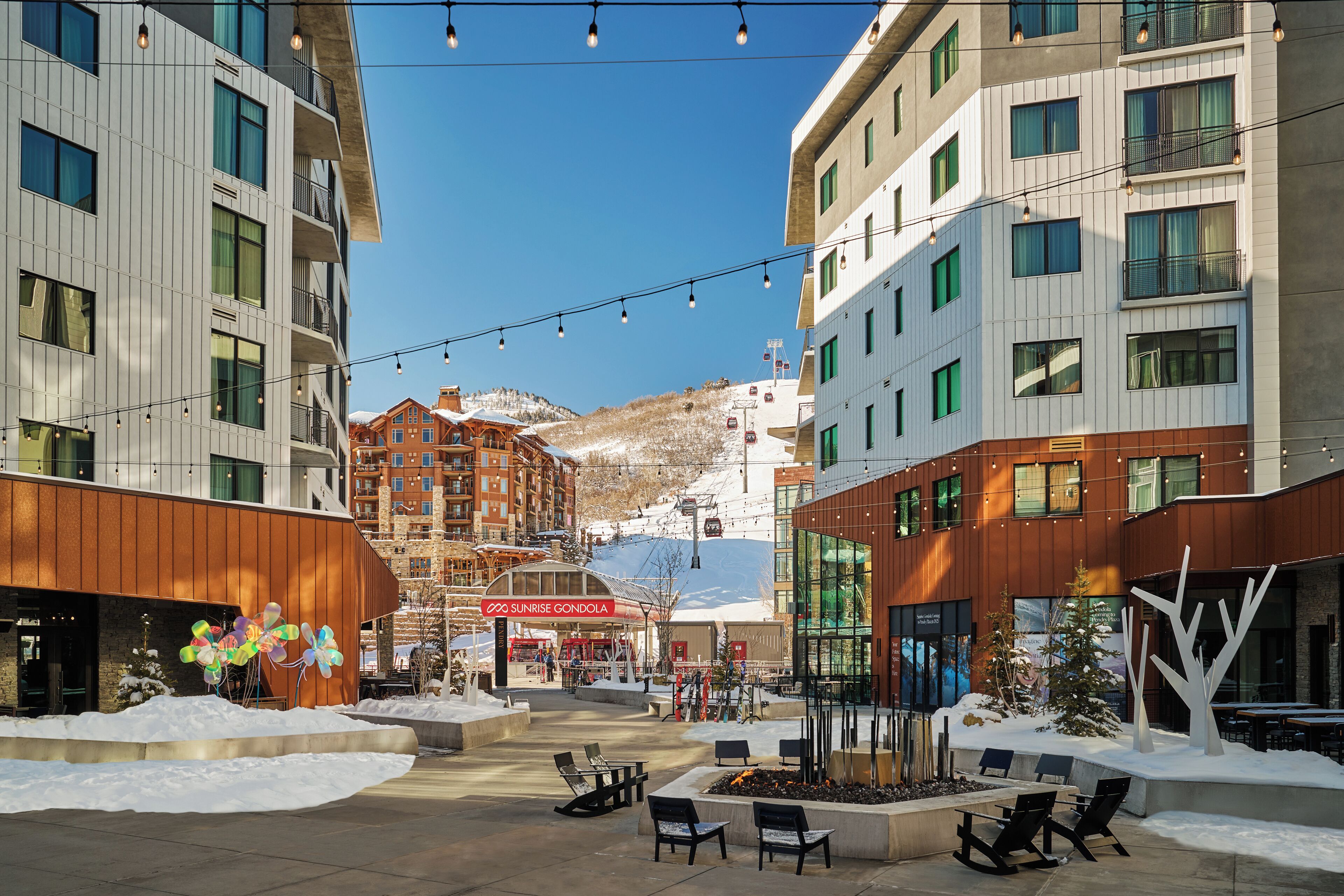 Una plaza nevada con mesas al aire libre, un telesilla al fondo y edificios modernos con balcones.
