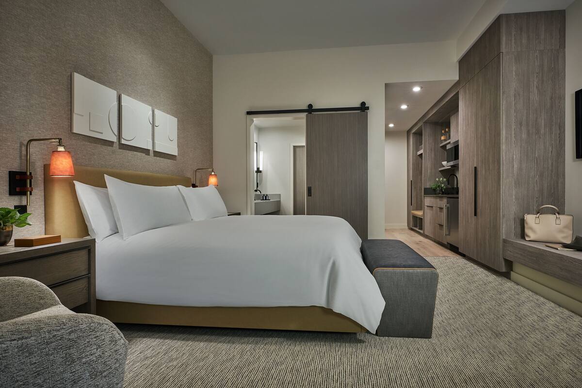 Une chambre moderne avec un grand lit, des tables de chevet, une commode, un banc, une armoire et un téléviseur fixé au mur.
