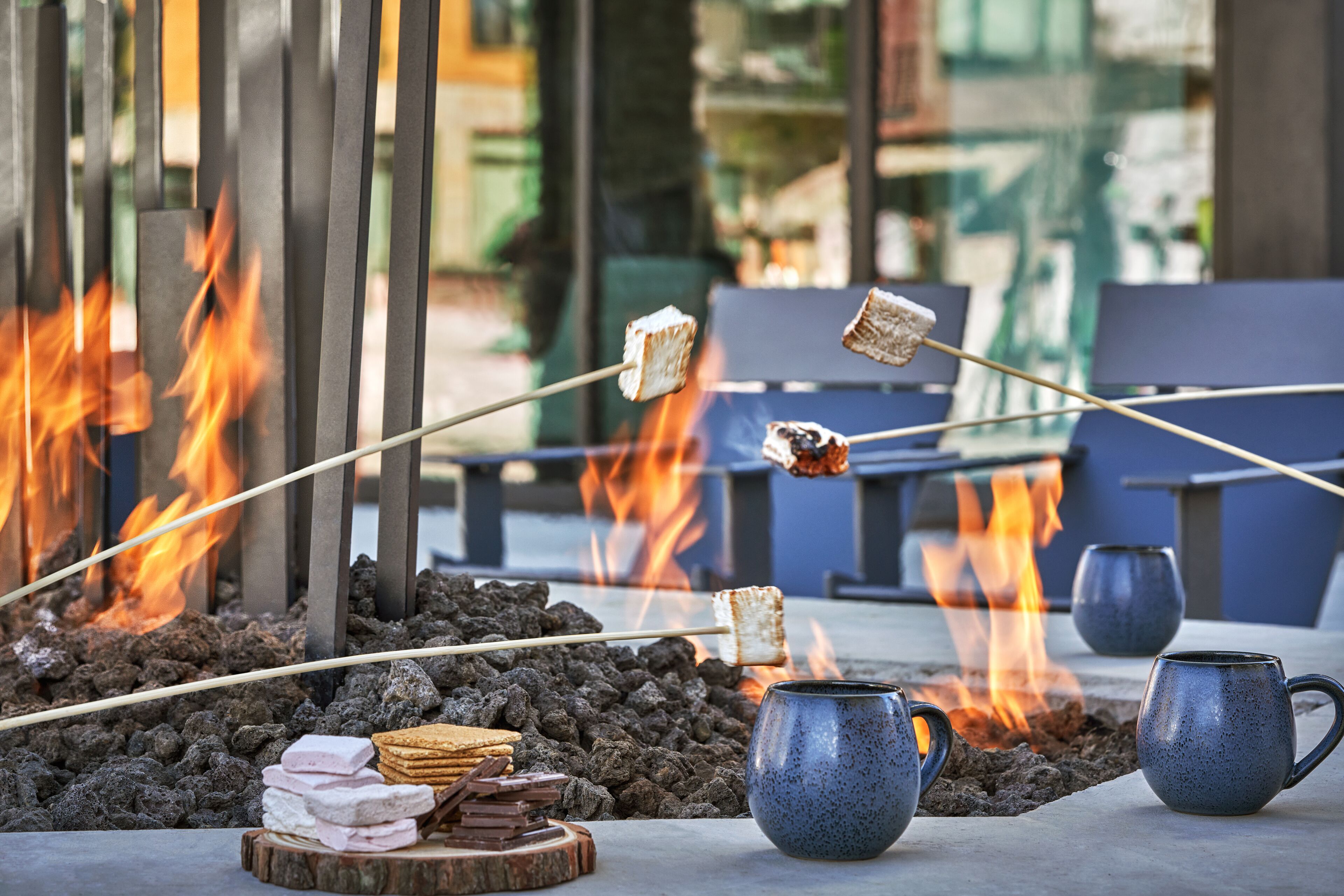 A fire pit with toasting marshmallows and a stack of chocolate bars, surrounded by seating and two blue mugs.