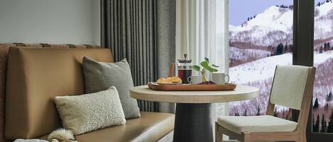 Une pièce chaleureuse avec un canapé marron, une table ronde sur laquelle est posée une assiette de pâtisseries, un fauteuil et une vue sur des collines enneigées.