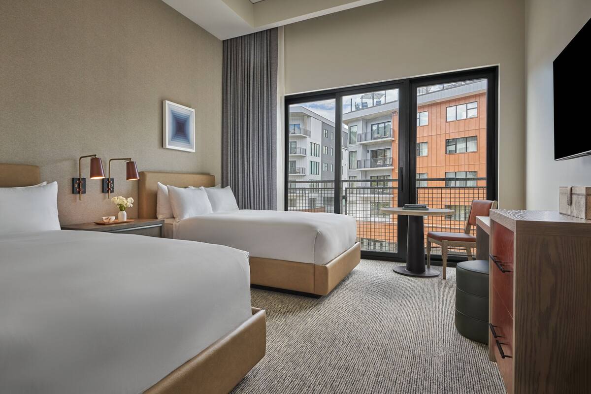Une chambre d’hôtel avec deux lits, un bureau, une chaise, une télévision et un balcon donnant sur des immeubles.