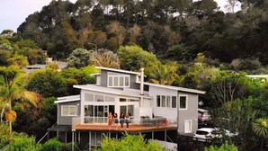 Uma casa moderna de dois andares com varanda, cercada por vegetação exuberante e colinas.