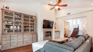 A spacious living room with a large grey sectional sofa, a wooden cabinet with glass doors, a flat-screen TV mounted above a fireplace, and a staircase leading to another level.
