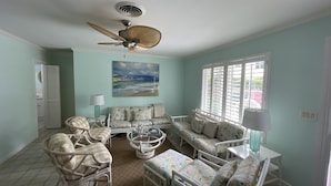 A living room with a ceiling fan, a painting of a beach, and a view through the window. - Oceanfront Beach Cottage with Private beach access in beautiful Juno Beach. (Juno Beach)