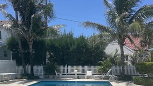 A rectangular swimming pool with clear blue water, surrounded by a white picket fence, palm trees, and a tiled roof house in the background. - Oceanfront Beach Cottage with Private beach access in beautiful Juno Beach. (Juno Beach)