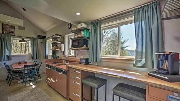 A modern kitchen with a central island, bar stools, a dining area with a table and chairs, and large windows with curtains. - Fire Pit + Gas Grill: Secluded Studio in Milford! (Milford)