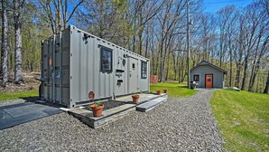 A compact RV with a grey exterior, a small attached shed, and a gravel driveway with potted plants. - Fire Pit + Gas Grill: Secluded Studio in Milford! (Milford)