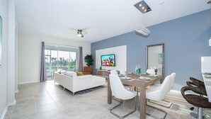 A modern living room with a white sofa, dining table, and chairs. There is a TV mounted on the wall and a large window with curtains. - DG1193 Champions Gate Condo 2Bedrooms 2Bathrooms Free Waterpark (Kissimmee)