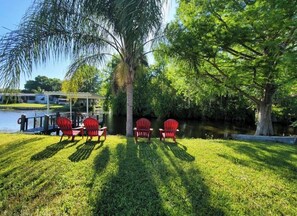 A grassy area with four red Adirondack chairs and a covered pavilion by a body of water.