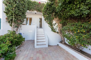 A white building with a staircase leading to a door, surrounded by greenery and plants.