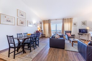 A modern living room with a dining area, a sofa, a TV, and a view of the sea.