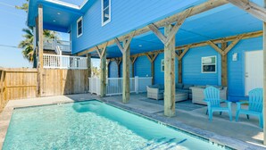 Una casa azul con piscina, terraza de madera y mobiliario exterior.