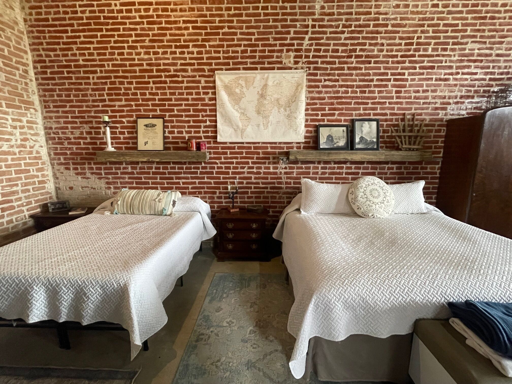 Una habitación con dos camas, una pared de ladrillo, un mapa del mundo y una estantería con fotos enmarcadas.