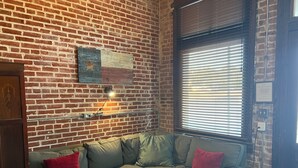 A cozy living room with a brick wall, a grey sectional sofa with red cushions, a floor lamp, and a large window with blinds. - Unique 1870's building with outdoor space to relax (McDade)