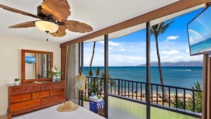 Une chambre avec une grande fenêtre donnant sur une plage, un ventilateur de plafond, une commode et un téléviseur.