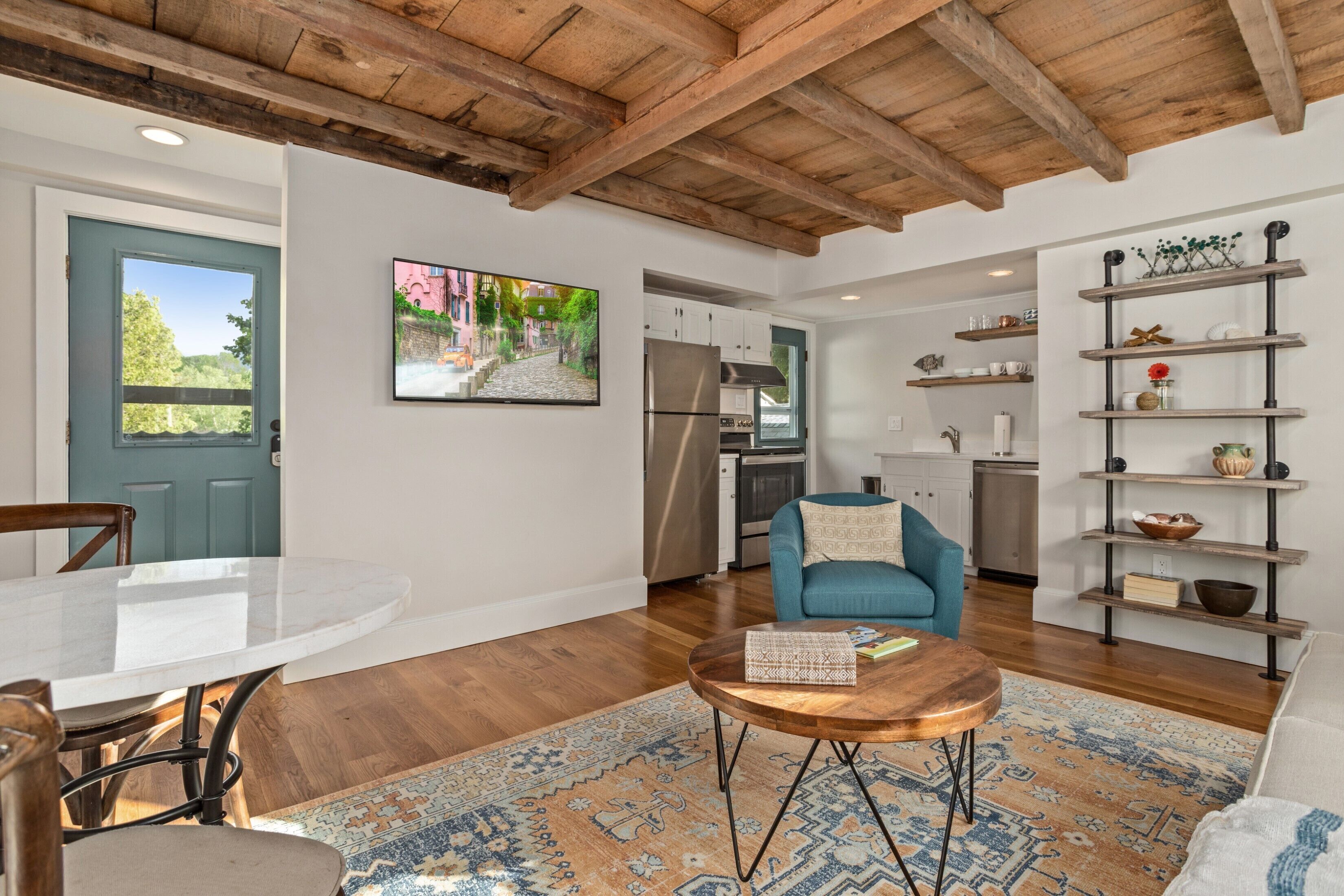 Un salon moderne avec un plafond en bois, une table à manger, un fauteuil bleu, une table basse et une cuisine en arrière-plan.
