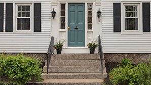 A white house with a green door, black shutters, and two potted plants on the front steps.