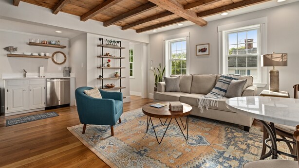 A spacious living room with a high ceiling, wooden beams, a sofa, a blue armchair, a coffee table, and a dining area with a table and chairs.