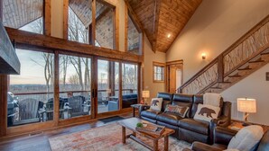 A spacious living room with a large window, a leather sofa, and a wooden staircase. - Happy Trails - Pet friendly Eagles Nest with view, hot tub, fire pit (Banner Elk)