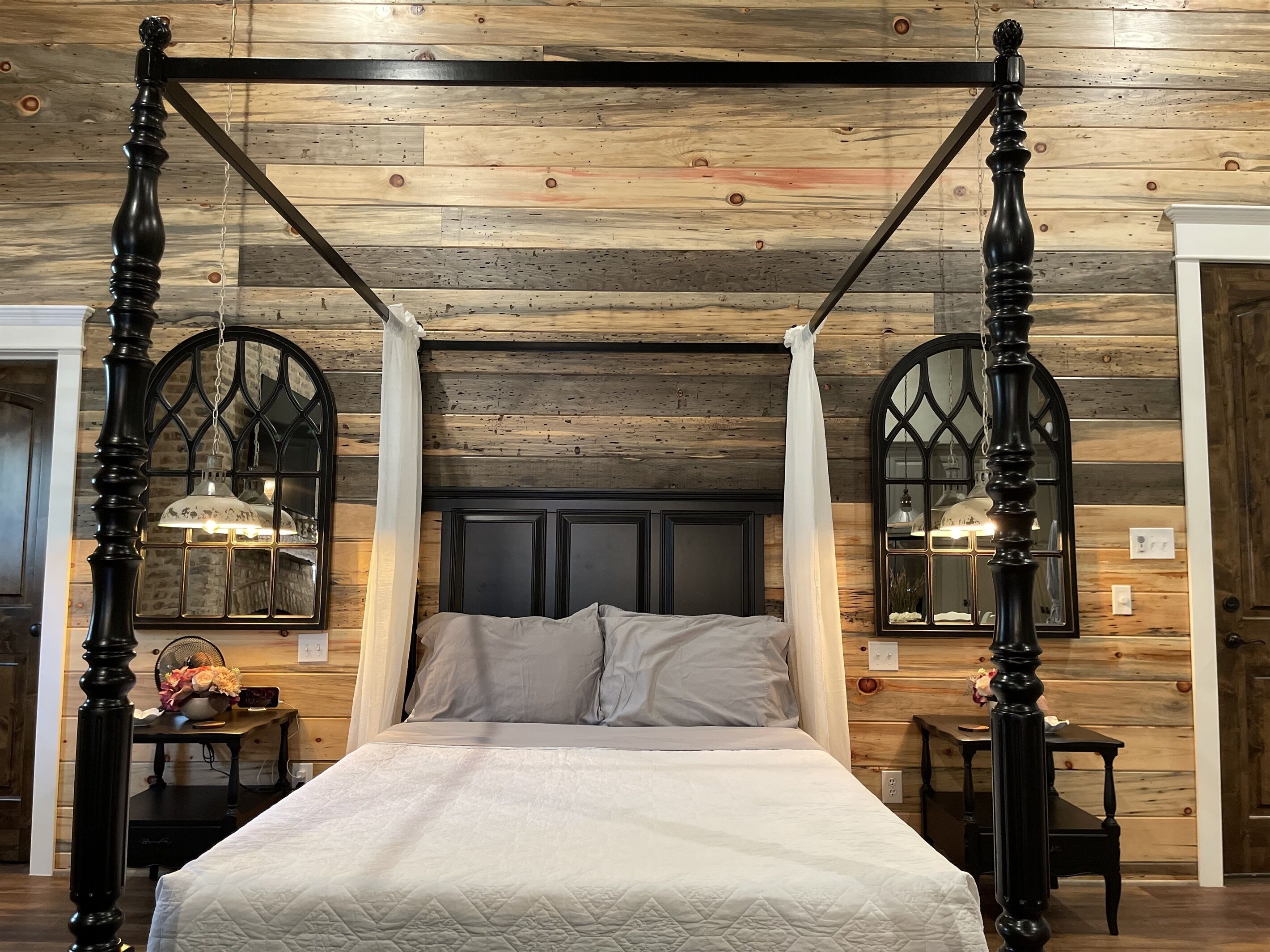 A four-poster bed with a quilted white bedspread, flanked by two wall-mounted lamps with glass shades, in a room with wooden paneling and a dark headboard.