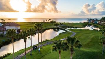 Un parcours de golf avec un plan d’eau, des palmiers et une vue sur l’océan au coucher du soleil.