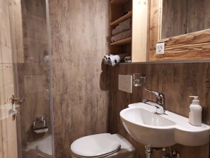 Ein Badezimmer mit Duschkabine aus Glas, Toilette, Waschbecken mit Seifenspender und Holzregalen über der Toilette.