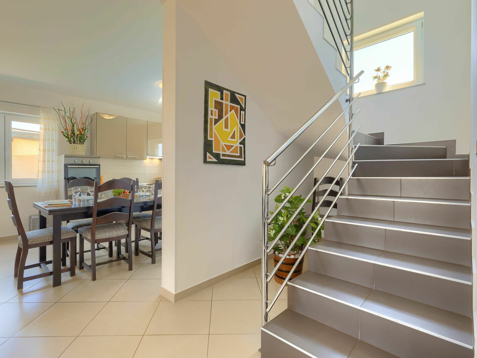 A modern kitchen with a dining area, a staircase with a metal railing, and a potted plant on the landing.