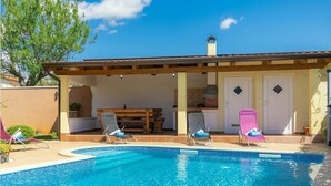 A pool area with lounge chairs, a covered patio, and a kitchen area.