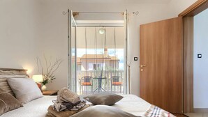 A bedroom with a bed, pillows, a bedside table with a lamp, a vase with flowers, and a view of a balcony with chairs and a table.