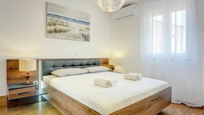 A spacious bedroom with a large bed, a wooden headboard, a painting of a beach scene, and a modern chandelier.