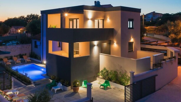 A modern two-story building with a swimming pool, outdoor seating, and well-lit windows.