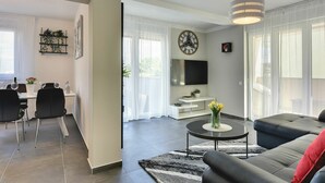 A modern living room with a sofa, a coffee table, and a TV. There is a dining area with chairs and a table.