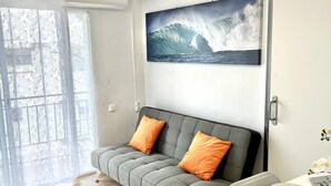 A modern living room with a grey sofa, orange cushions, a dining table with plates and cutlery, and a large wave painting on the wall.
