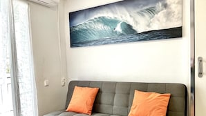 A grey sofa with two orange cushions, a large wave painting on the wall, and a door with a handle.