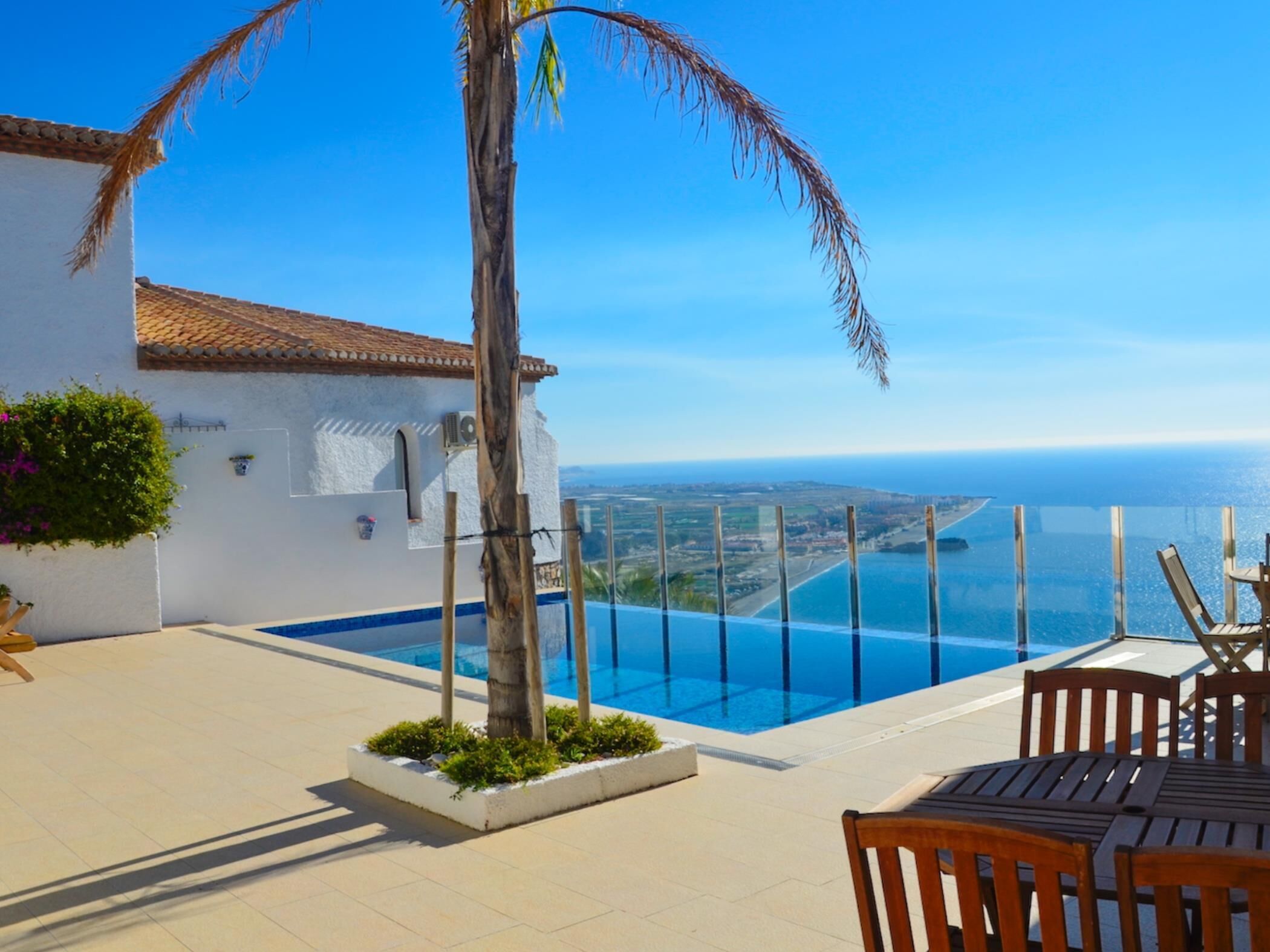 Un espace aménagé au bord de la piscine offrant une vue dégagée sur l’océan, avec un palmier et un bâtiment blanc doté d’un toit en tuiles.