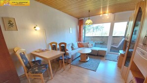 A cozy living room with a wooden ceiling, a dining table with chairs, a sofa, and a large window with curtains.