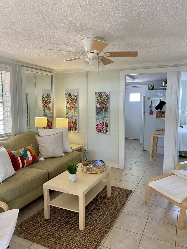 A living room with a sofa, coffee table, and a ceiling fan. There are paintings on the wall and a kitchen area in the background.