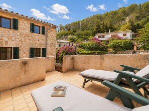 A stone building with a balcony, outdoor seating, and a view of a hillside with trees and houses.